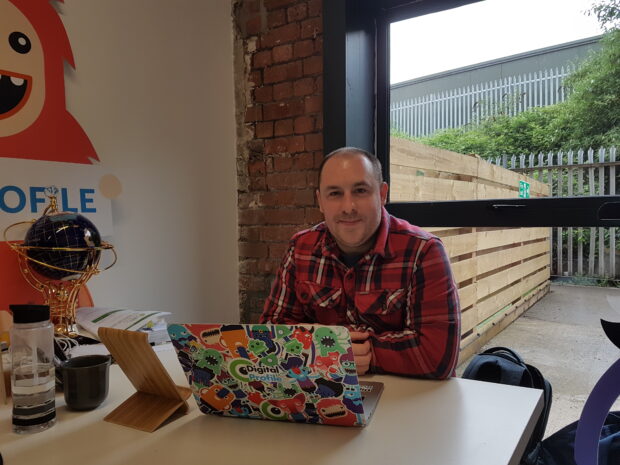 A picture of Daniel Lewis CEO of Digital Profile smiling at the camera sat whilst sat at a desk behind his laptop. 