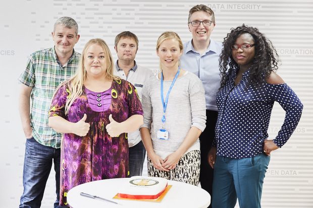 6 members of the team standing behind the llama cake. 