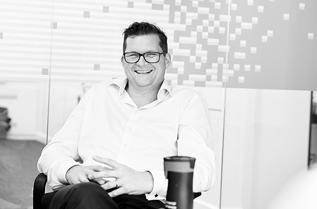 A black and white image of Ross Maude, Director of Digital sitting on a chair and smiling. 