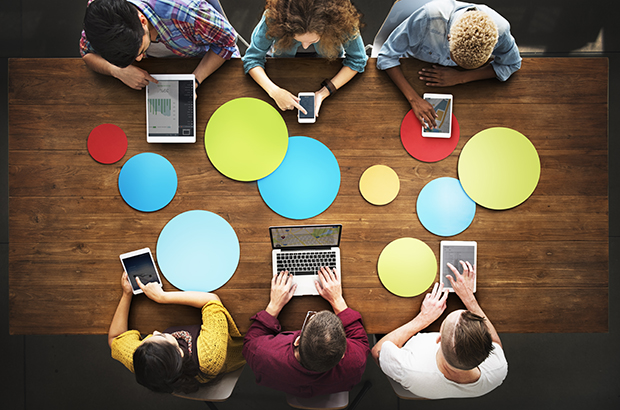 A group of young people sitting around a table using laptops, tablets and mobile phones