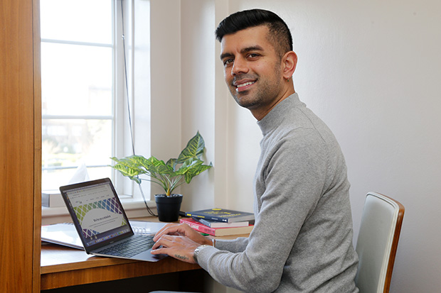 A person smiling at the camera whilst working on a laptop at a desk.