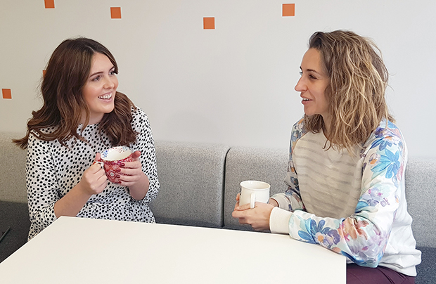 Two females smiling and chatting holding cups of coffee.