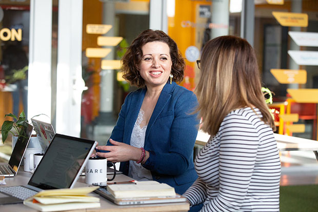 Two people chatting at a desk full of books, coffee and a laptop. One of the people is Zara Nanu, CEO and co-founder of Gapsquare.