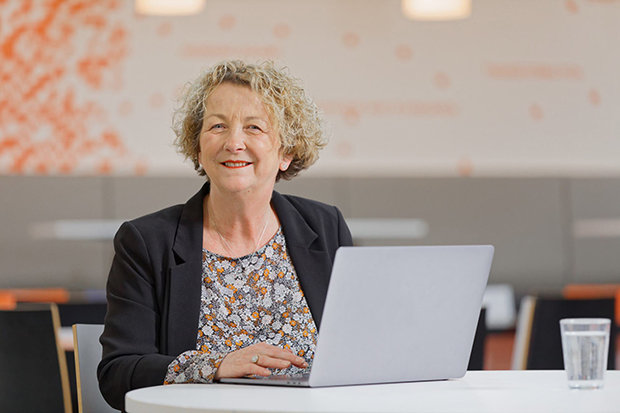 A lady sat at a desk behind a laptop and smiling at the camera.