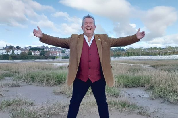 A man standing outdoors wearing warm clothing and holding his arms out wide and smiling.