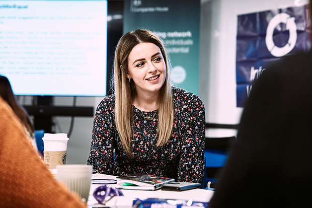 A lady looking to the side and smiling whilst sat at a desk in a conference room.