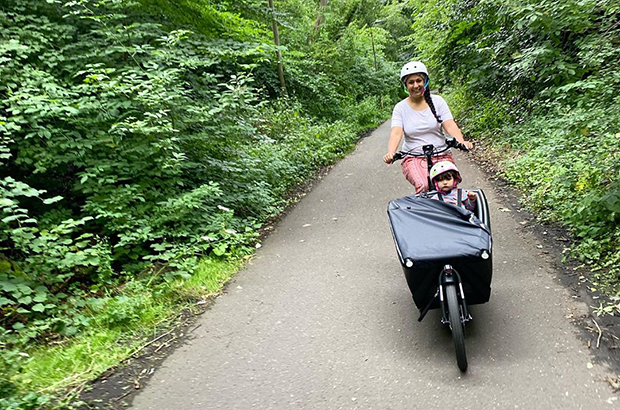 A woman, wearing a helmet, driving a cargo bike in a country lane. 