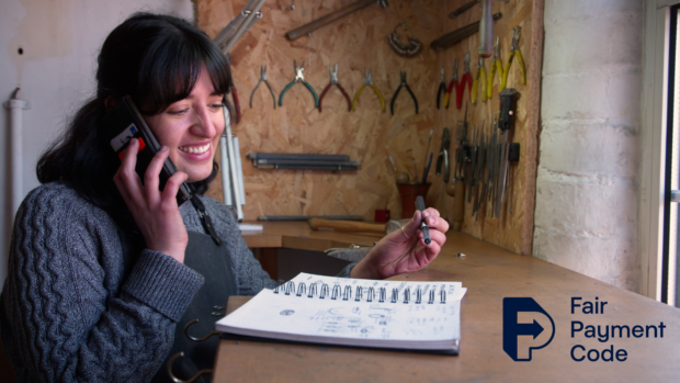 A woman is sat in her workshop, talking on the phone with a notebook in front of her