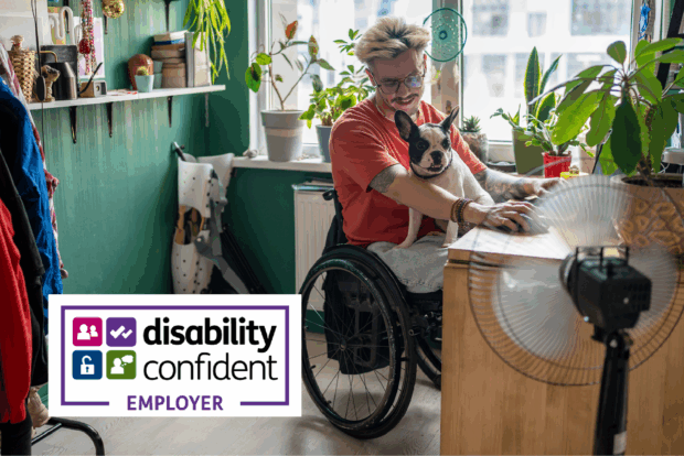 A young man in a wheelchair is sitting in a home working space with a dog on his lap.