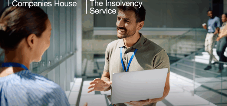 Two people in an office setting are talking to each other whilst holding a laptop.